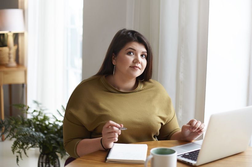 Žena s nadváhou sedící u stolu a pracující s počítačem