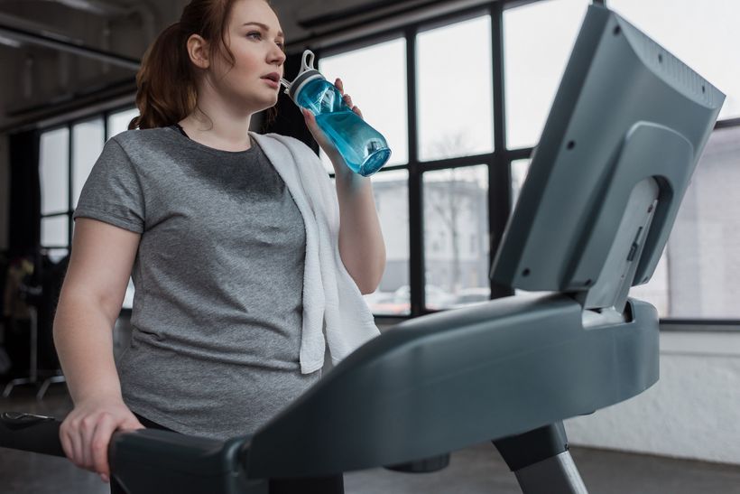 Spocená žena pijící vodu z flašky po běhání na běhacím stroji.